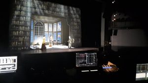 View of a stage during a technical rehearsal.  Various monitors and light boards are visible in the foreground.  Three actors in period costume stand on a raked stage with projected scenery around it.