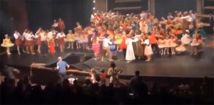 Image of a stage with many children and teenagers.   Part of the stage has collapsed and many have fallen through the hole.