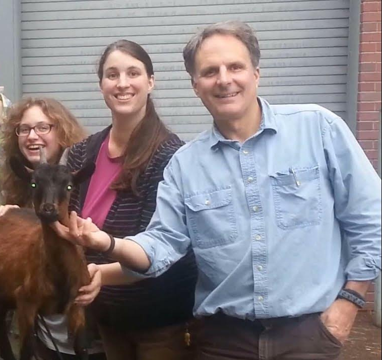 Bob Bruemmer is shown wearing a blue shirt outside of a loading dock with two students.  He is petting a baby goat.