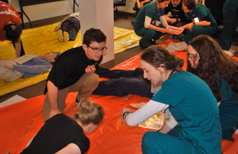 Various students are crouched around an orange tarp.  Some are lying prone as if injured.  Some are wearing nurses scrubs.  The woman in scrubs in the foreground looks intently at her watch.