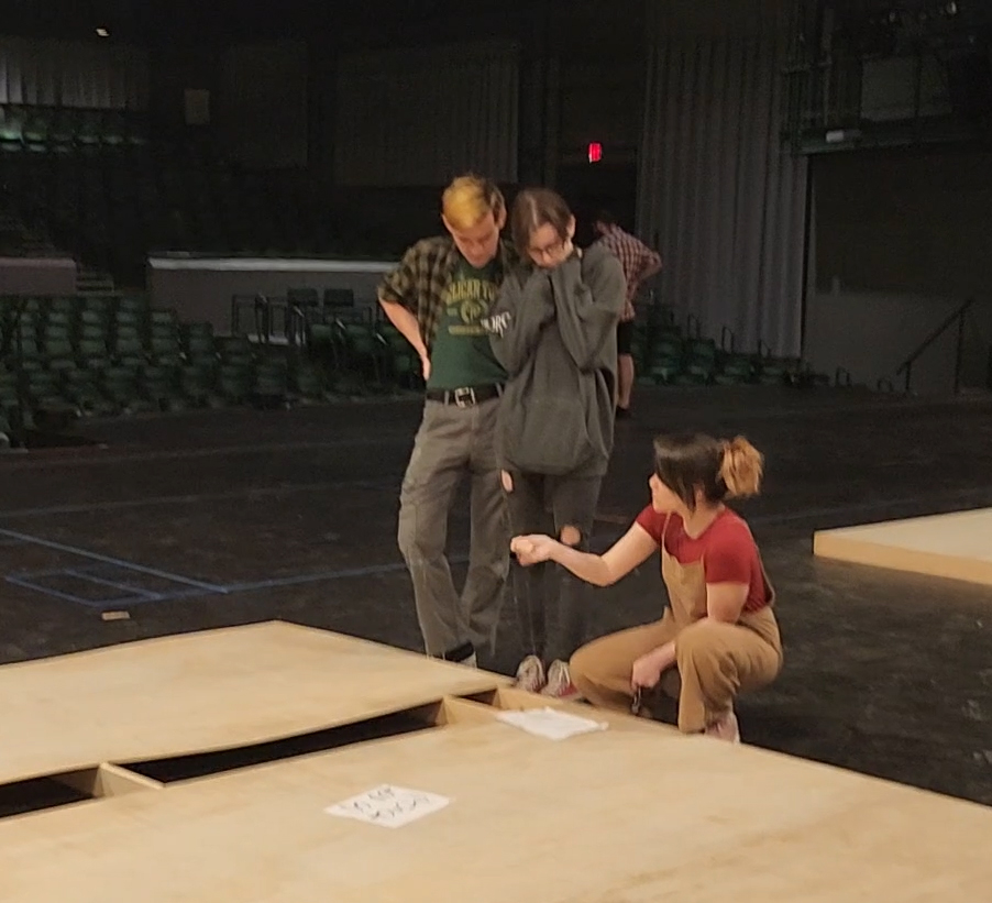 Heather Manfredi is crouched on the floor next to a large piece of scenery on a stage.  Audience seating is visible in the background.  She is speaking to two college students who are listening attentively.