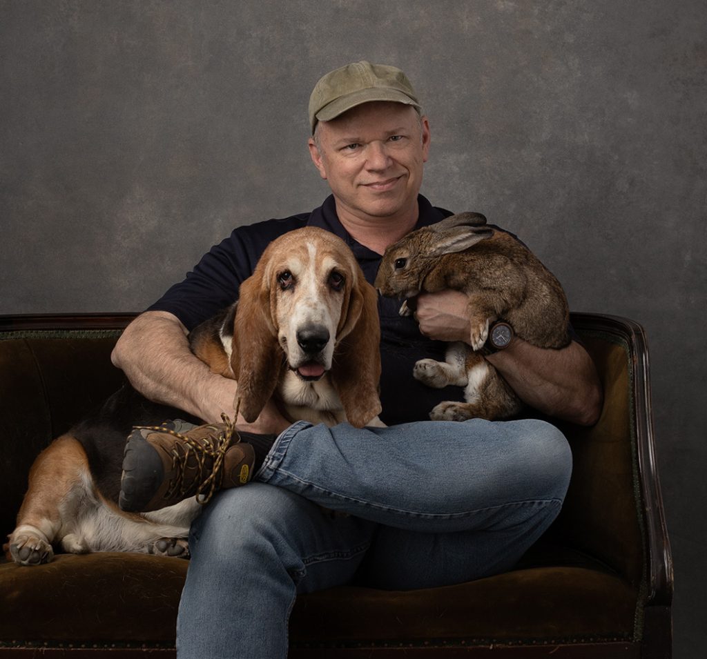 Matt Kizer is shown seated wearing a khaki ball cap with one arm around a basset hound and the other holding a brown rabbit.  All three are smiling.