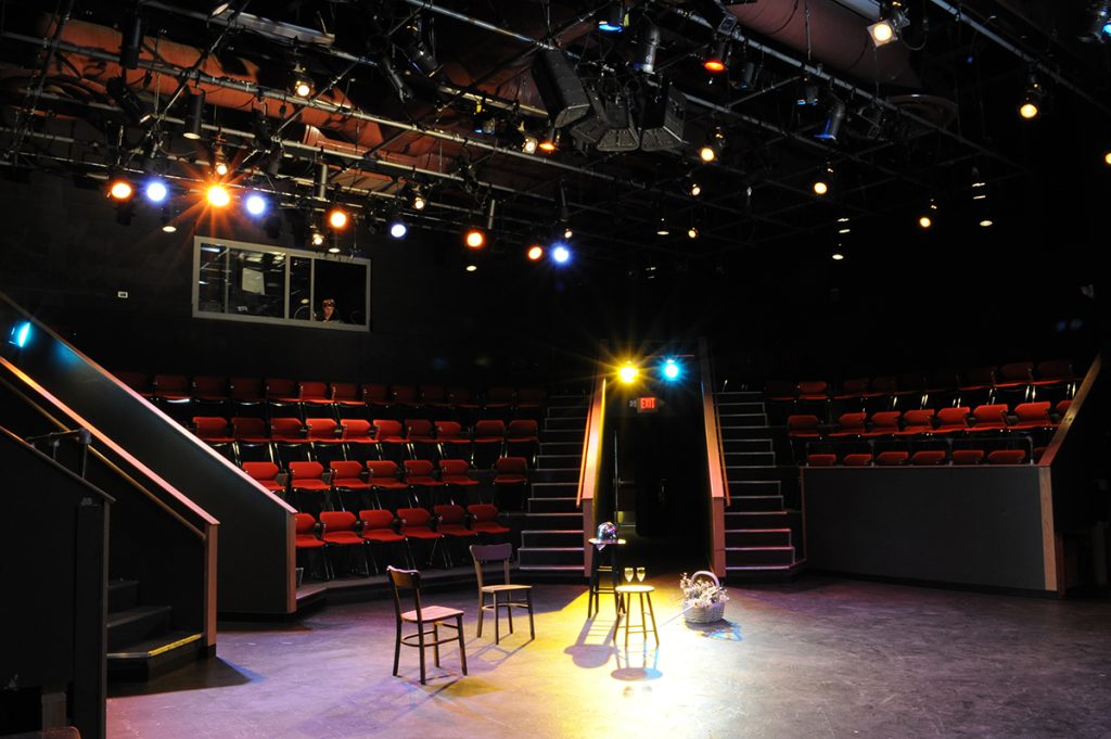 The view from the stage of a three-quarter thrust performance space.  The control booth is visible over the audience space which is made up of comfortable looking red seats.